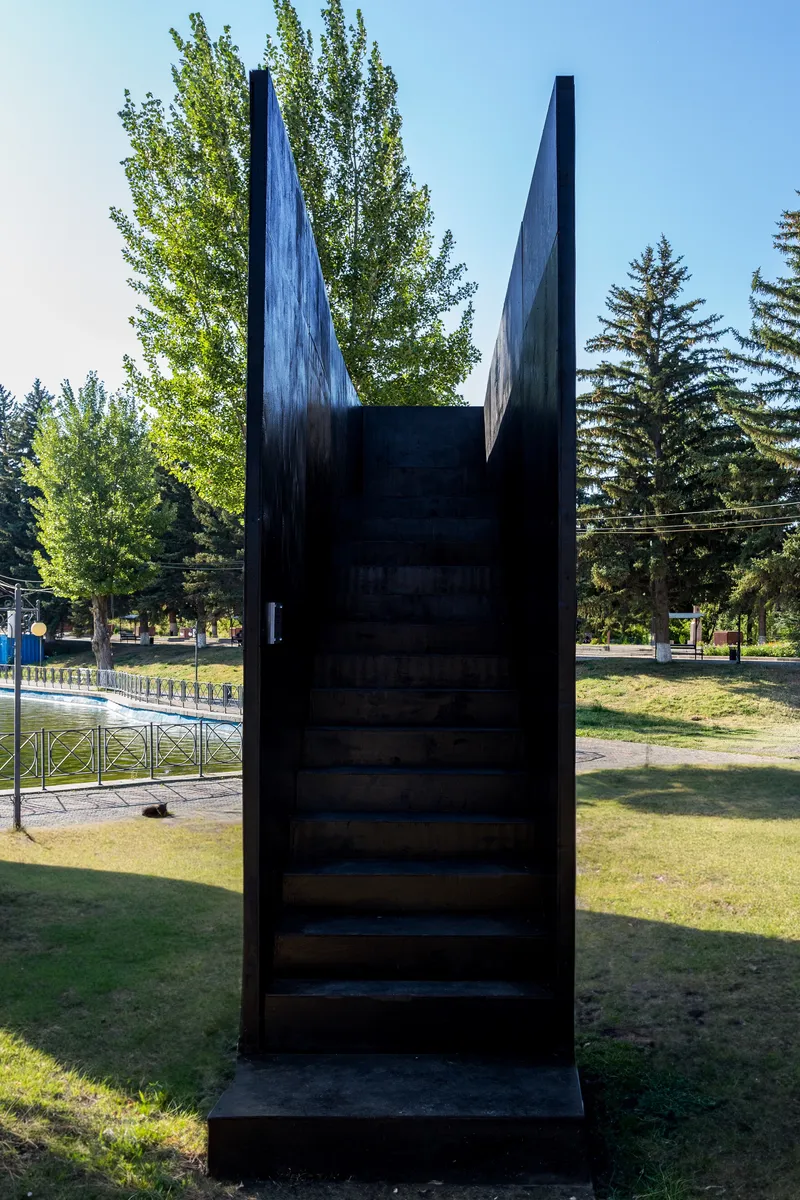 Dark metal staircase leading up between two angled mirrored panels surrounded by park landscape