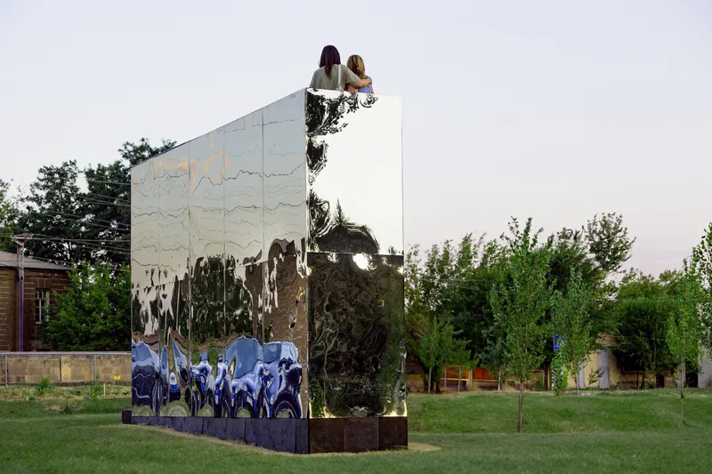 Mirrored panel installation reflecting two figures atop a dark plinth within a green lawn at dusk