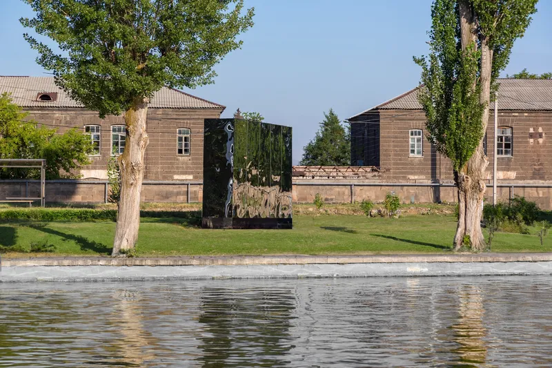 Reflective panel sculpture on grass with a shallow pool in the foreground and residential buildings behind