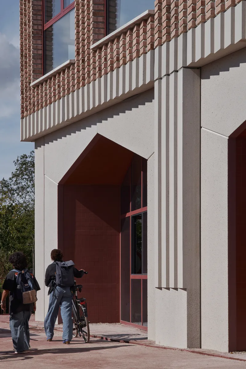 Close-up of arched maroon entrance portal with vertical fluted precast cladding and people approaching with bicycles
