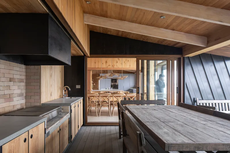 Interior dining area with timber ceiling beams and recycled wood table looking through to kitchen beneath dark range hood