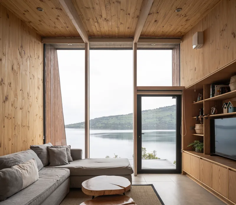 Living room with timber-clad walls and ceiling featuring floor-to-ceiling glazing framing a view across the lake to hills