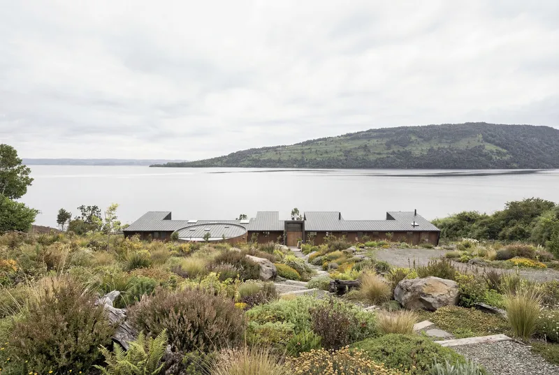 Low-profile metal-roofed volumes nestled into a rocky hillside with native planting overlooking a calm lake and forested peninsula