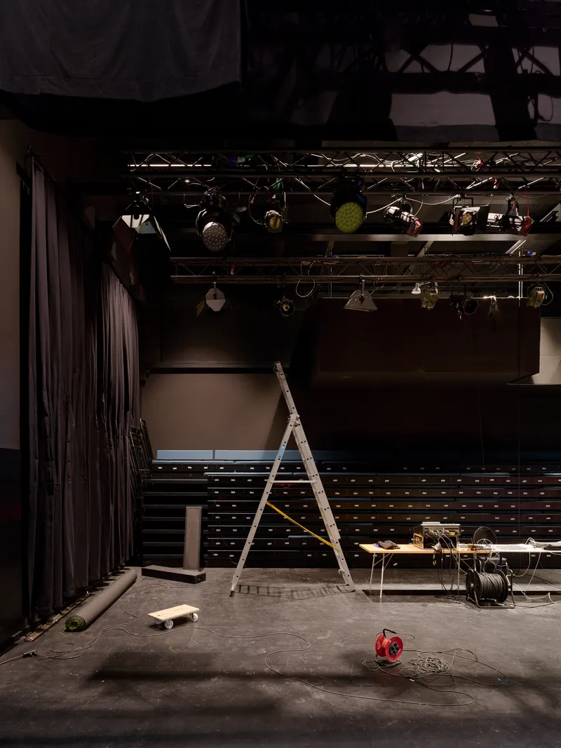 Stage area with lighting rig overhead and ladder resting among scattered equipment on concrete floor