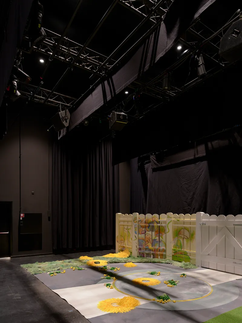 Black box theater space with exposed truss rigging and a stage floor painted with yellow and green patterns