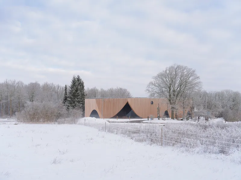 Wood-clad volume with arched openings set in a snow-covered field beneath bare winter trees
