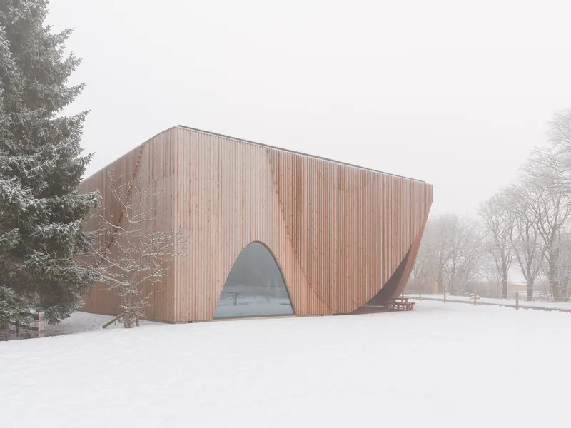 Curved timber facade with arched glass entry framed by evergreen and deciduous trees in winter
