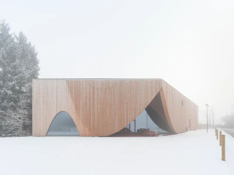 Vertical timber-clad facade with arched cutouts set in a snow-covered field under heavy mist