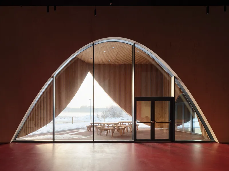 Arched glazed opening framing a wood-lined porch and snowy landscape beyond a red interior floor