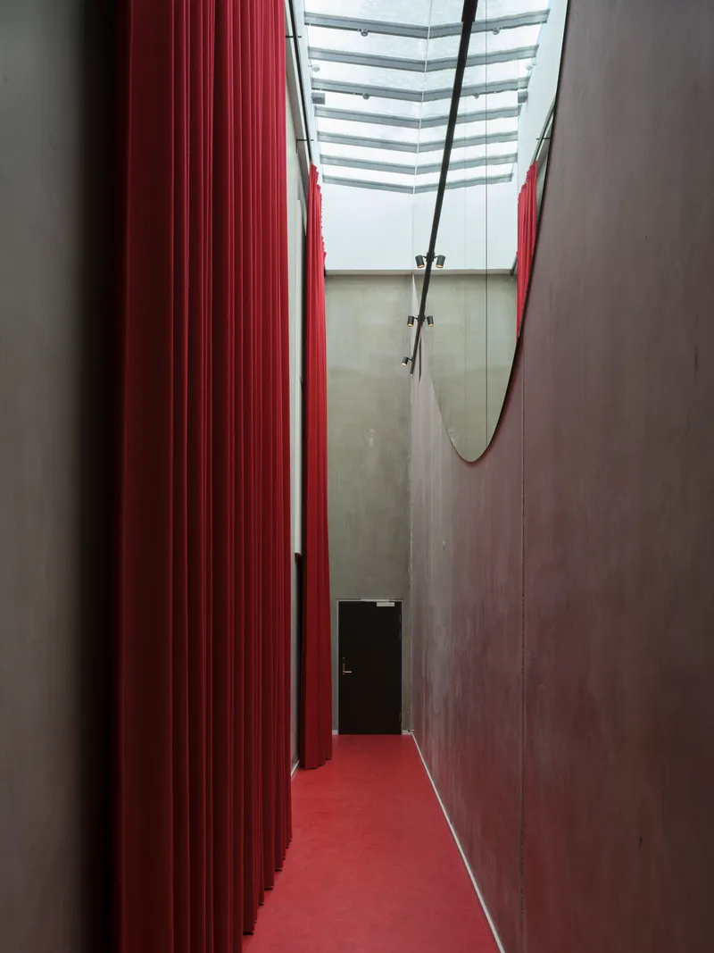 Narrow corridor with red ribbed wall and matching floor beneath a skylight with suspended trapeze