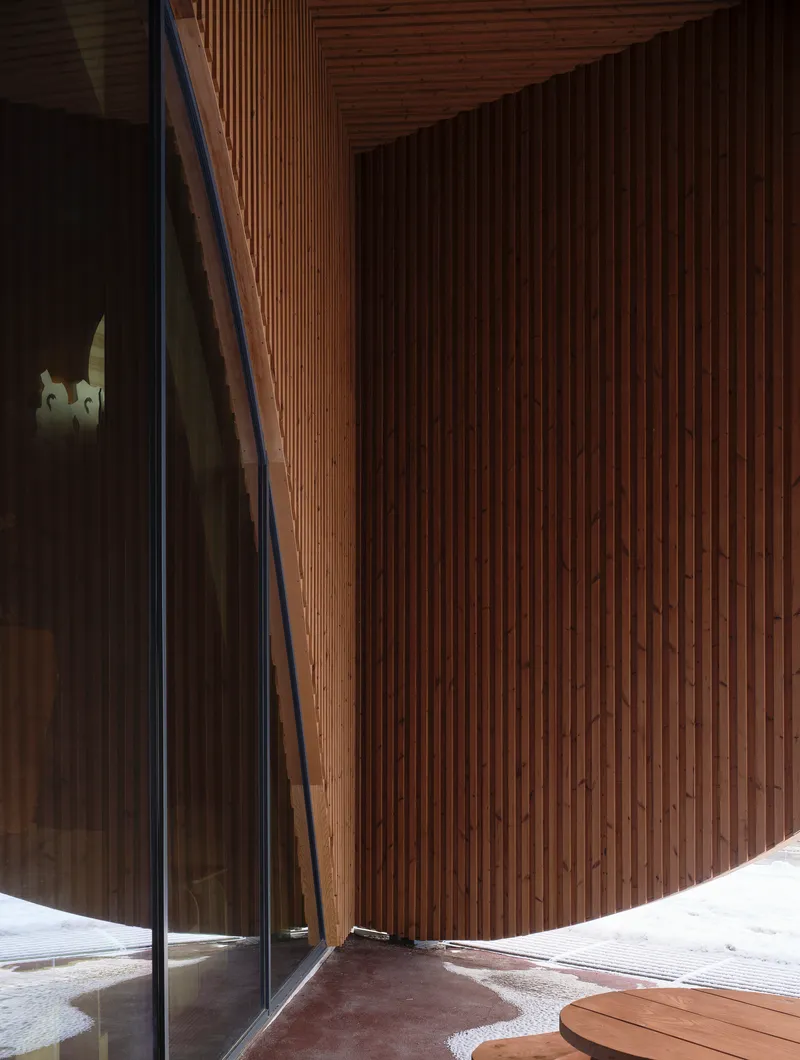 Curved vertical timber slat wall meeting a frameless glass corner at the entrance threshold