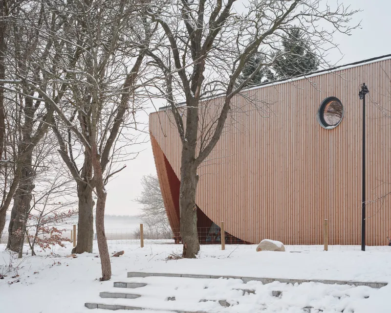 Vertical timber facade with circular window and diagonal entrance beneath bare winter trees