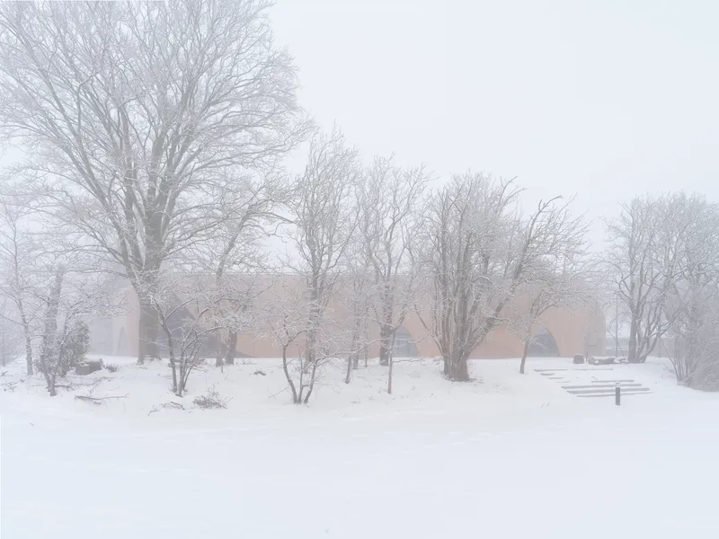 Distant view of the low-slung beige brick structure amid bare winter trees in heavy snow and fog