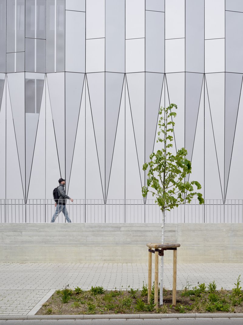White paneled facade with geometric relief pattern behind a young street tree and walking pedestrian