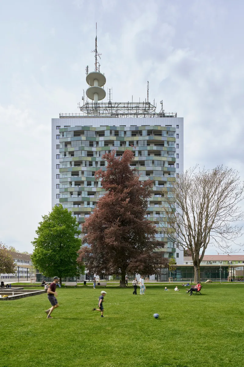 White tower with multicoloured balconies and rooftop antenna array behind a park lawn with children playing