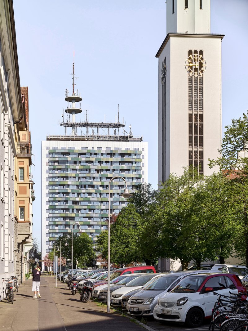 Tower with multicoloured balconies and rooftop antenna seen between a church bell tower and a street parking lot