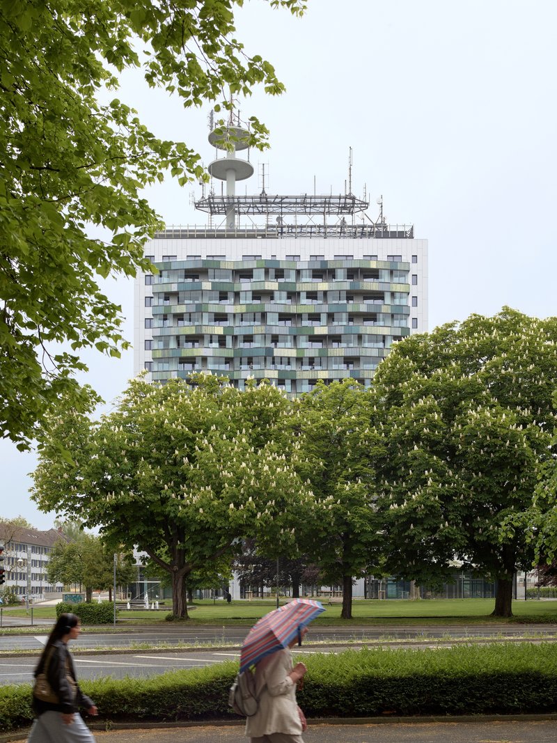 Residential tower with rooftop antenna framed by park trees with a person holding an umbrella in foreground