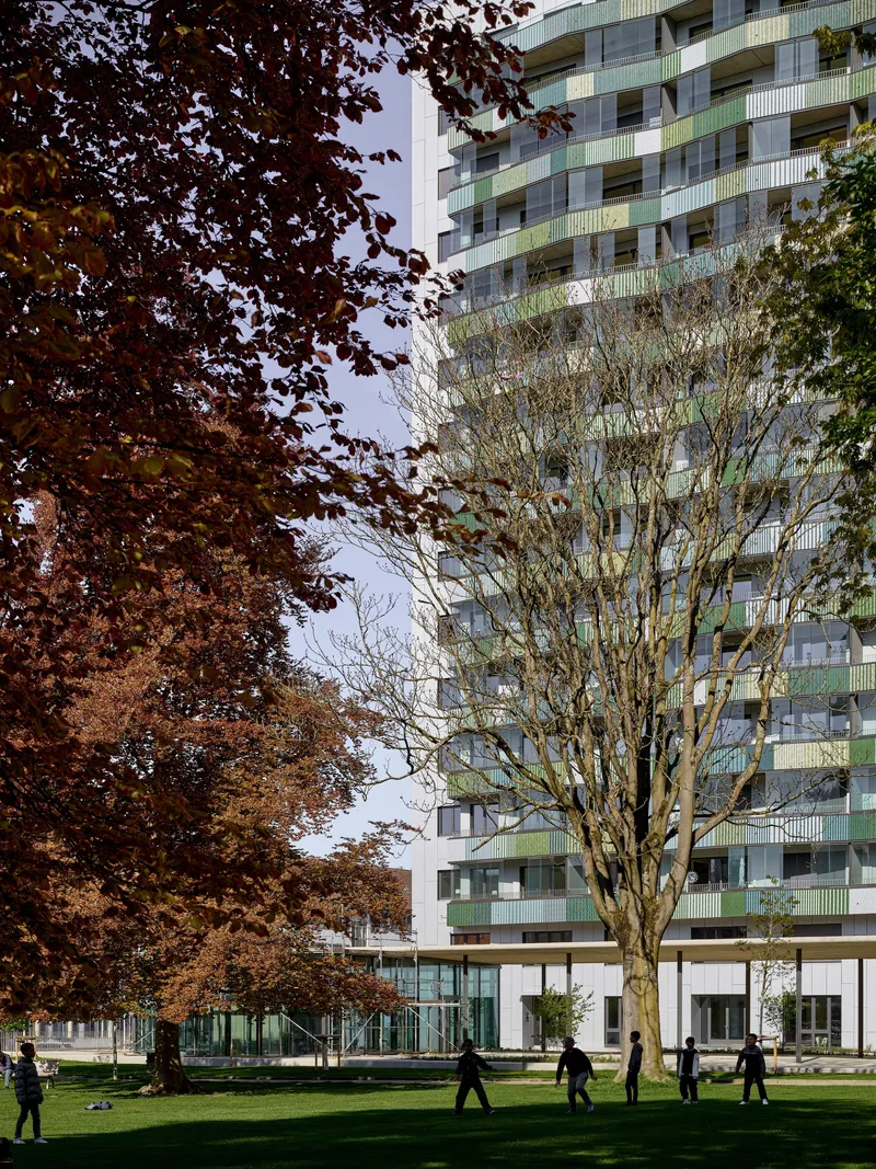 Facade with glass balconies and green railings partially obscured by autumn trees in the park below