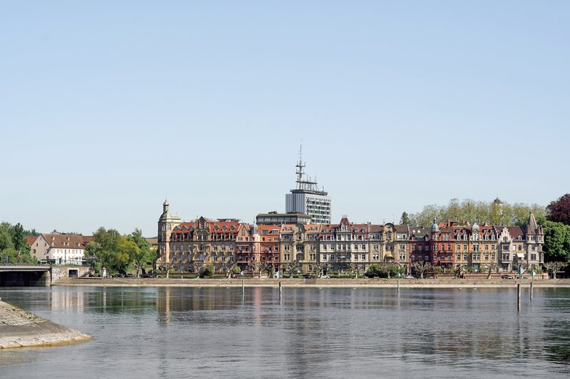 Riverfront view of residential blocks with a glass tower rising above the roofline under spring sky