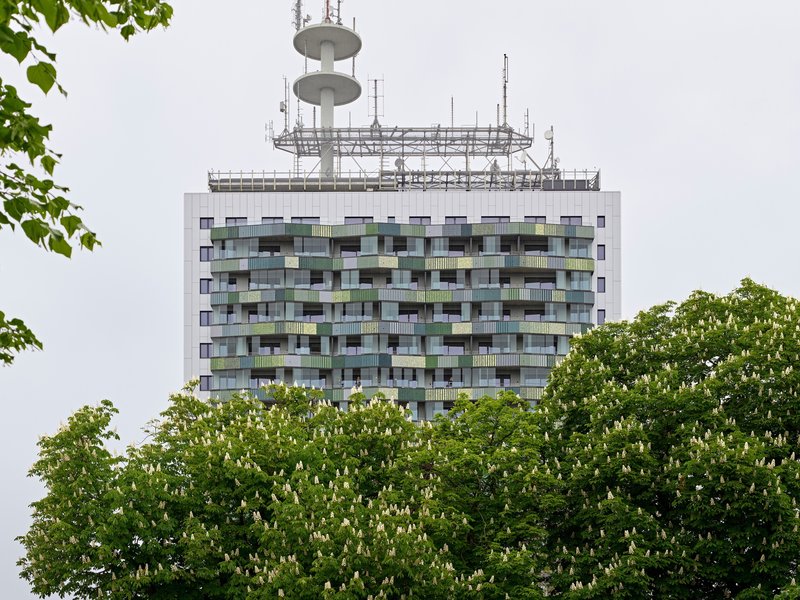 Residential tower with patchwork balcony facade and rooftop telecommunications mast framed by green foliage