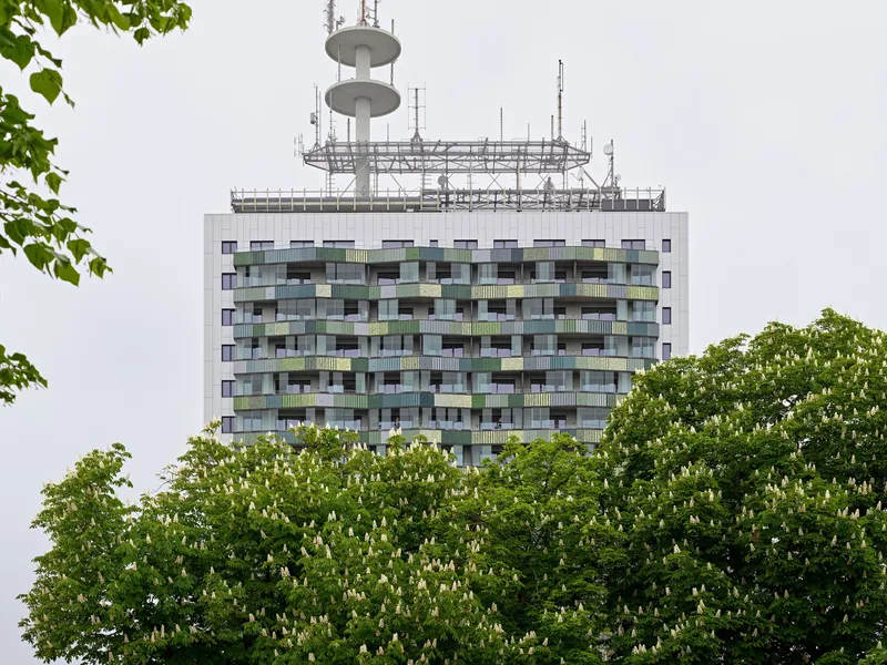 Residential tower with patchwork balcony facade and rooftop telecommunications mast framed by green foliage