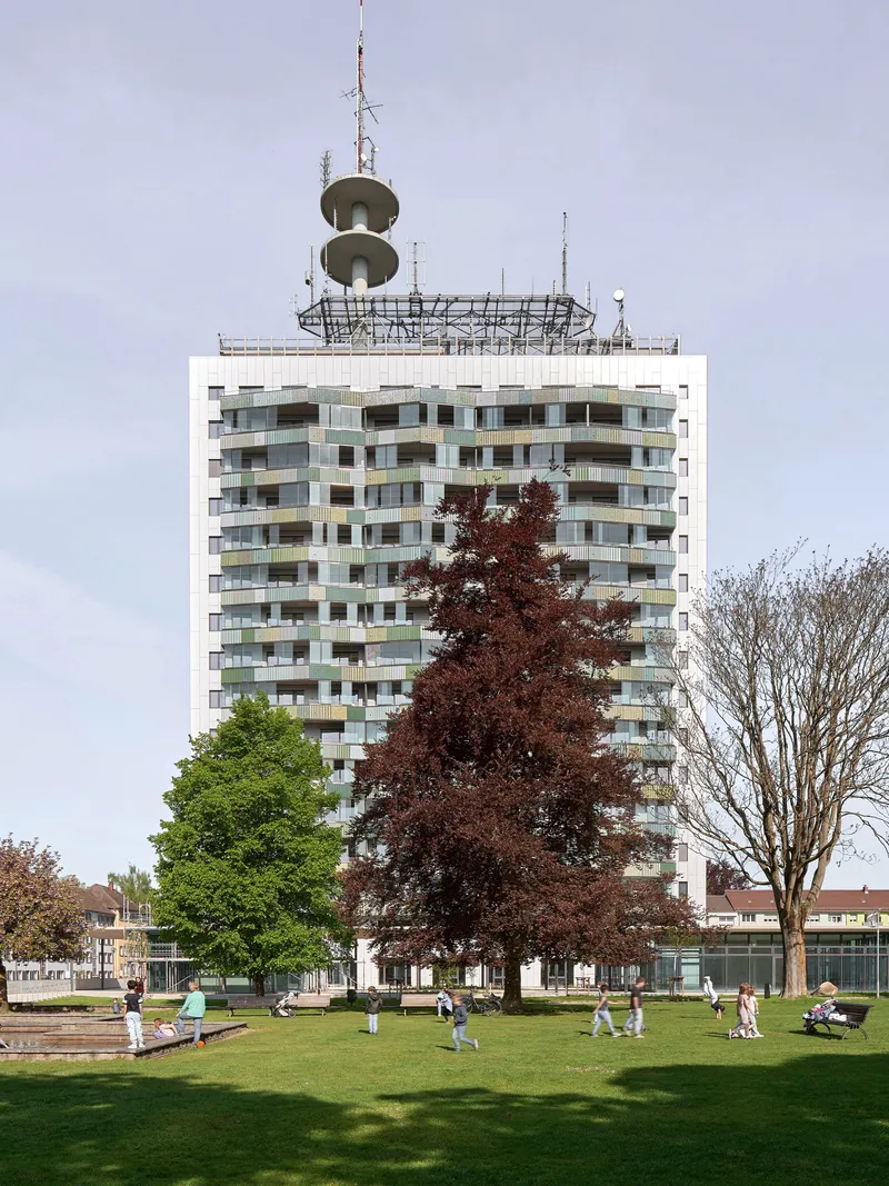 Tower with multicolored balcony grid rising above a public lawn with spring trees and scattered visitors