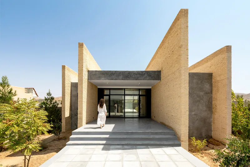 Covered entrance with pale stone steps and person in white approaching glazed doors under concrete beam