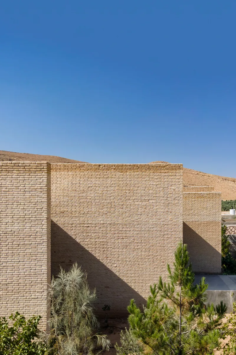 Staggered buff-colored brick walls forming angular volumes with desert shrubs in the foreground
