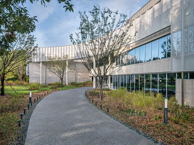 Paved walkway flanked by planted beds and young trees leading toward the translucent curtain wall facade