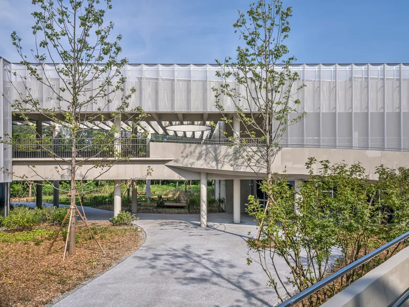 Elevated bridge connecting building volumes over a landscaped ground plane with young trees and dappled shadows