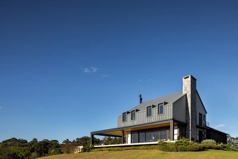Two-storey gabled house with stone chimney and covered terrace set on a grassy hillside under clear blue sky