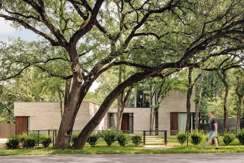 Street view of the horizontally banded stone facade with timber panels beneath mature oak trees