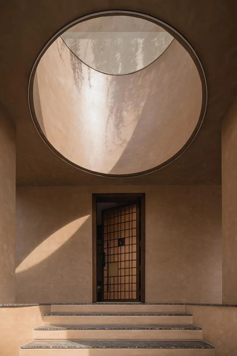 Entry steps leading to gridded door beneath circular skylight with curved plaster walls in afternoon light