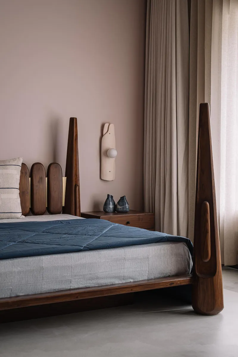 Bedroom corner with sculpted walnut bed frame, wall-mounted light fixture and floor-length curtains in soft daylight