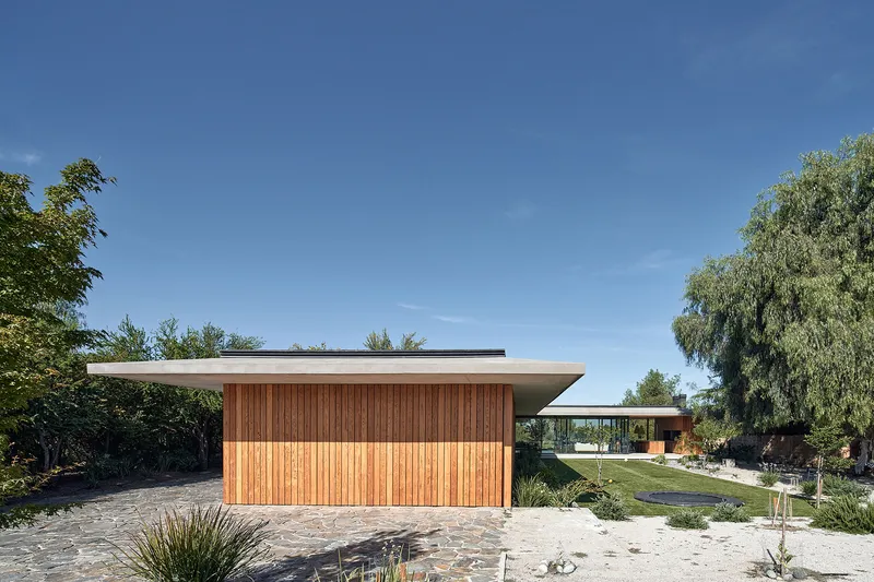 Street view of the timber-clad volume with extended roof plane and stone paving under clear skies