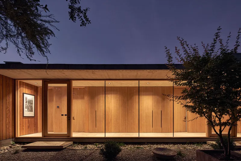 Illuminated timber facade with flat roof and glazed entry corridor at twilight beside planted gravel beds