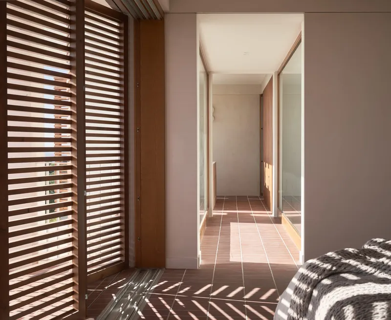 Corridor with timber louvered doors filtering sunlight into geometric shadow patterns on terracotta tile and pink walls