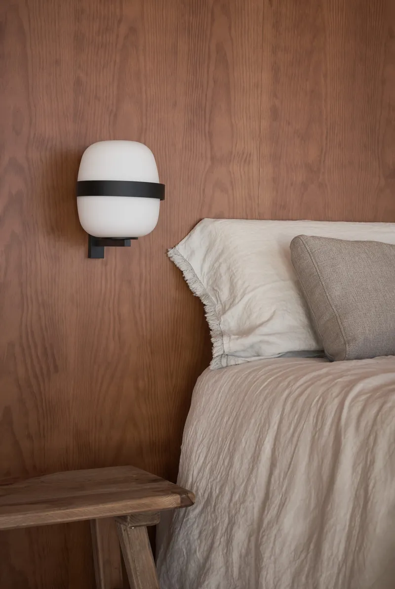 Bedroom wall clad in vertical wood grain panels with a wall-mounted white sconce above linen bedding