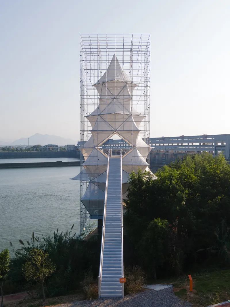 Tiered white tower enclosed by an outer steel scaffolding frame with a ramped pedestrian bridge at dusk