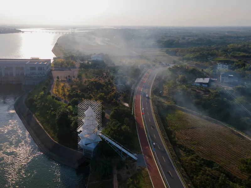 Aerial view of the scaffolded tower on a riverside peninsula beside a red bicycle path in morning mist