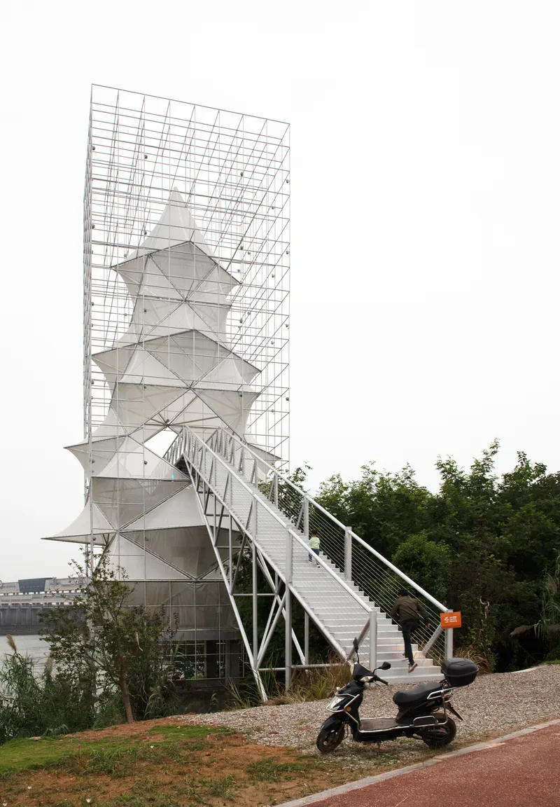 Side view of the steel scaffolding frame enclosing the stacked pyramidal volumes with a scooter parked below