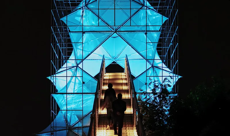 Nighttime view looking up through the illuminated blue glass panels and steel bracing with silhouetted figures on the stairs
