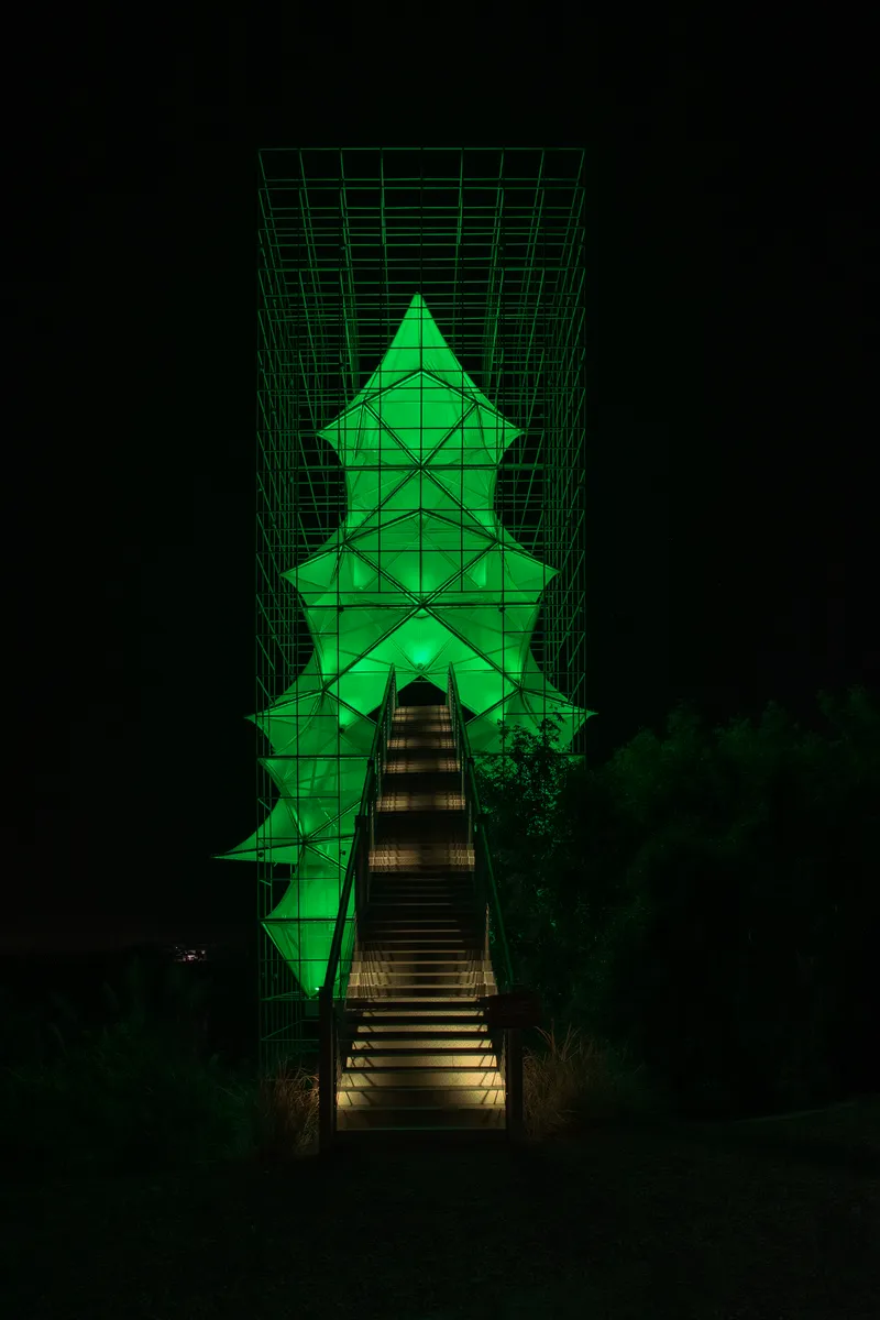 Illuminated pyramidal tower with green lighting and structural grid framing accessed by a central timber staircase at night