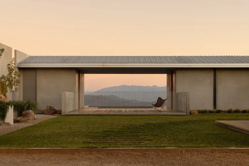 Garden view of the linear pavilion with corrugated metal roof and glazed opening framing distant mountains at dusk