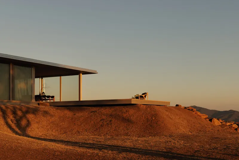 Glass-walled pavilion emerging from the hillside with outdoor terrace and chair at golden hour