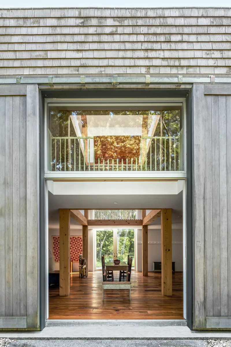 Exterior opening in weathered timber cladding revealing double-height interior with timber columns and balcony at dusk