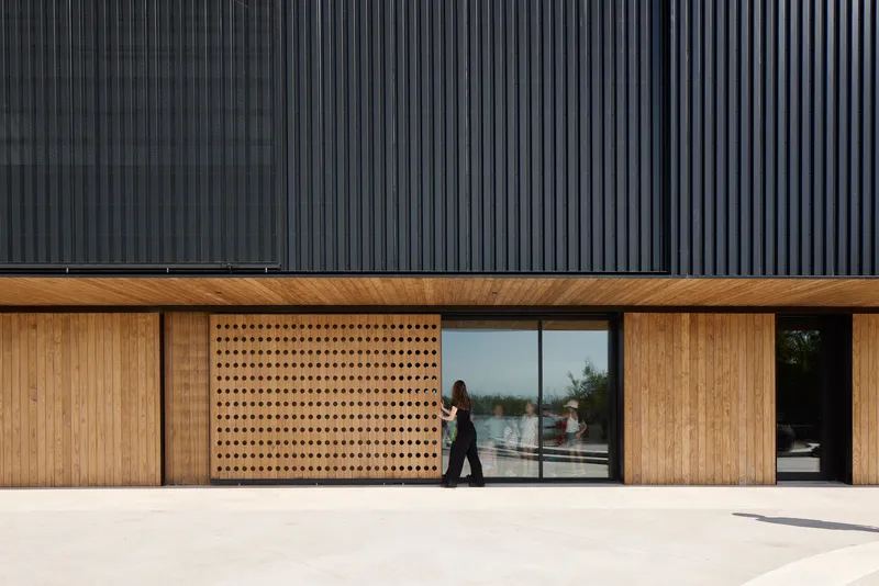Ground-level facade with perforated timber panels and glazed openings beneath dark corrugated metal cladding as a person walks past