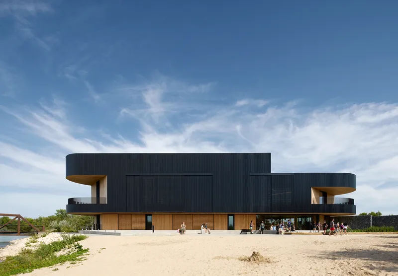 Two-story volume with black corrugated cladding cantilevering over timber base, set in sandy terrain with visitors gathering outside