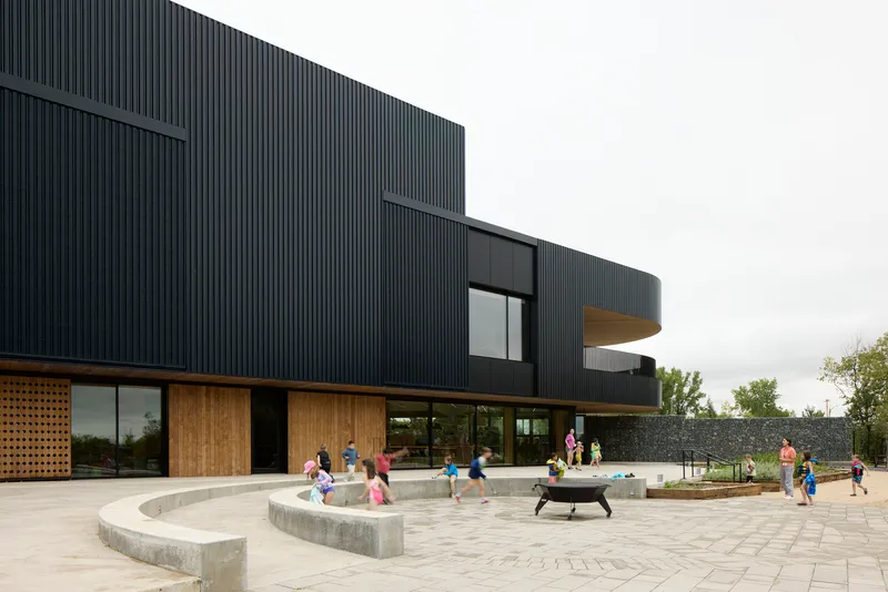 Curved corrugated facade with timber canopy overhangs and paved courtyard where children play around concrete benches