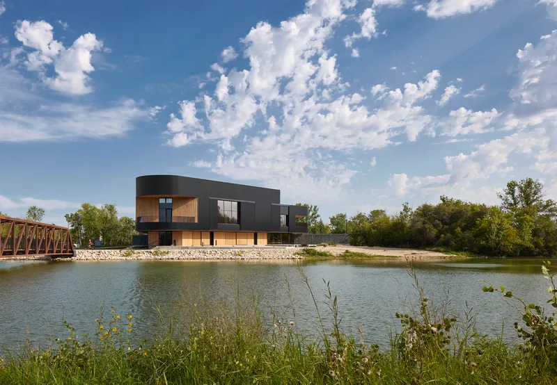 Two-story volume with curved metal cladding overlooking a pond with wildflowers and a truss bridge beyond